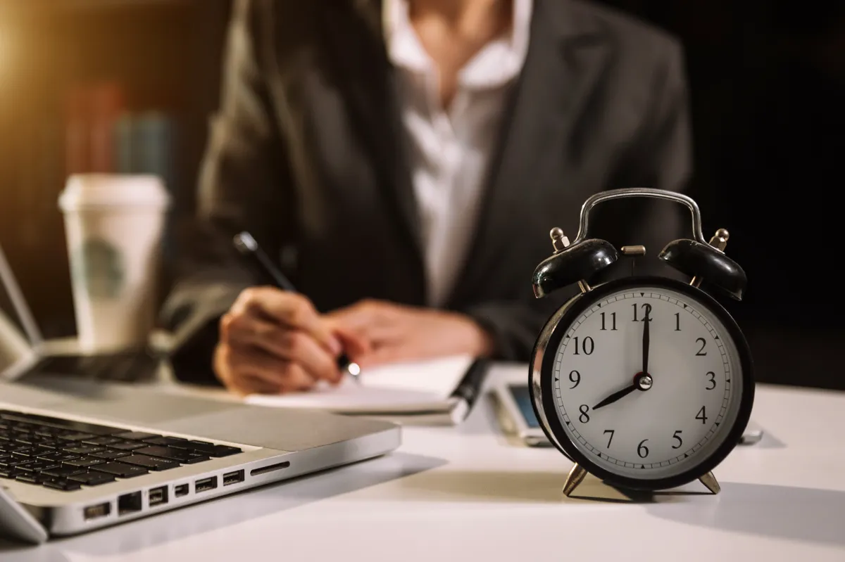 An alarm clock on a desk beside a professional taking notes, representing workplace time management