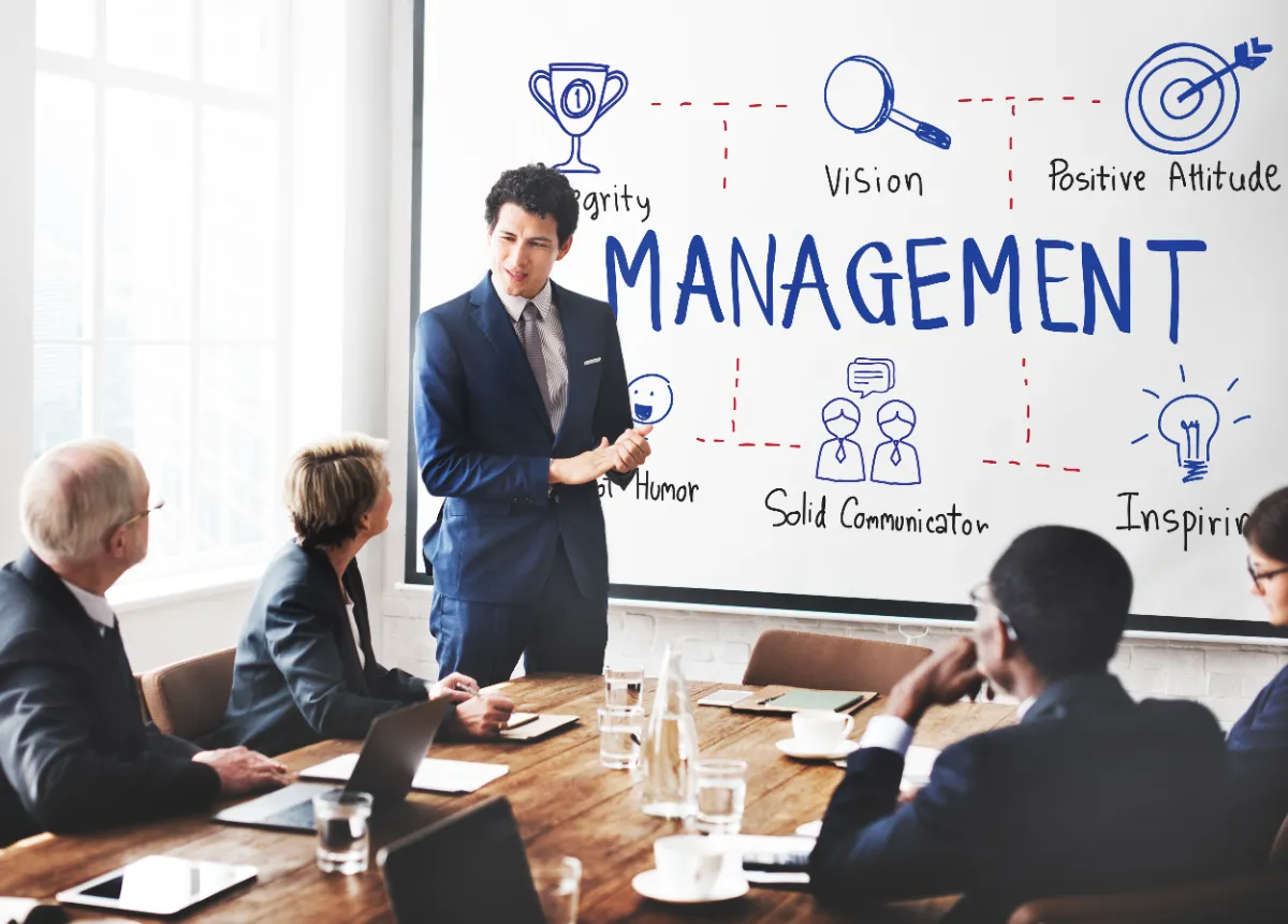 A presenter leading a boardroom discussion on management principles, with a diagram of vision, communication and leadership traits