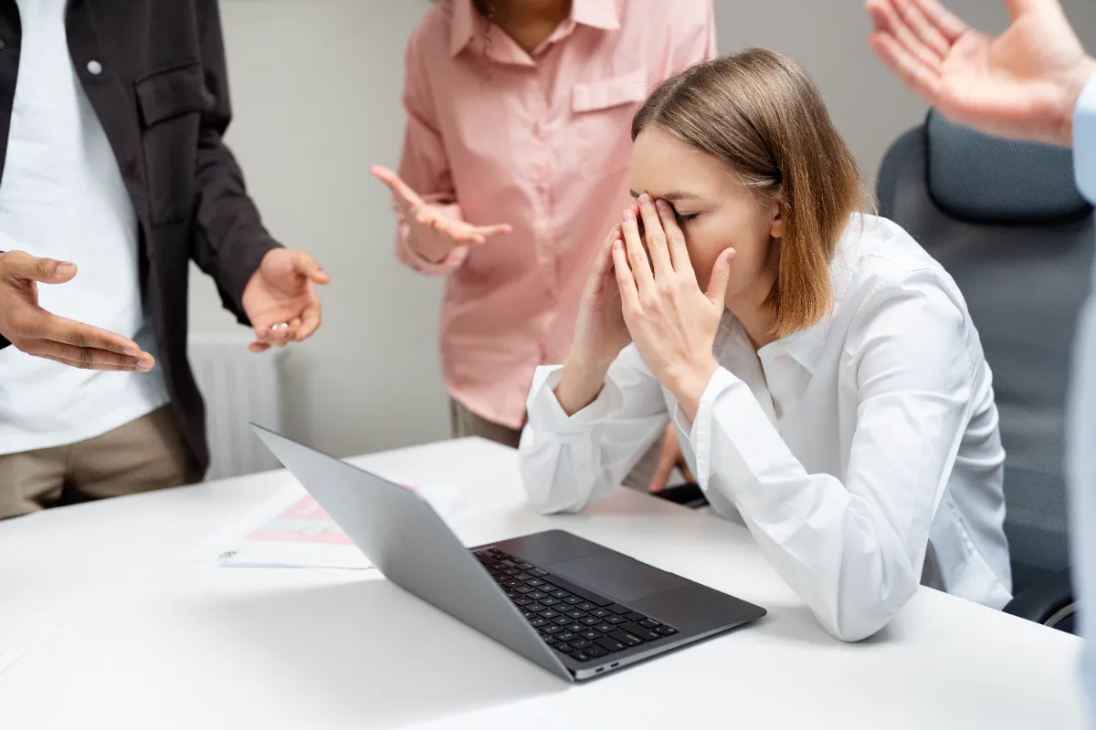 A distressed employee at her desk while coworkers gesture accusingly around her, illustrating workplace harassment